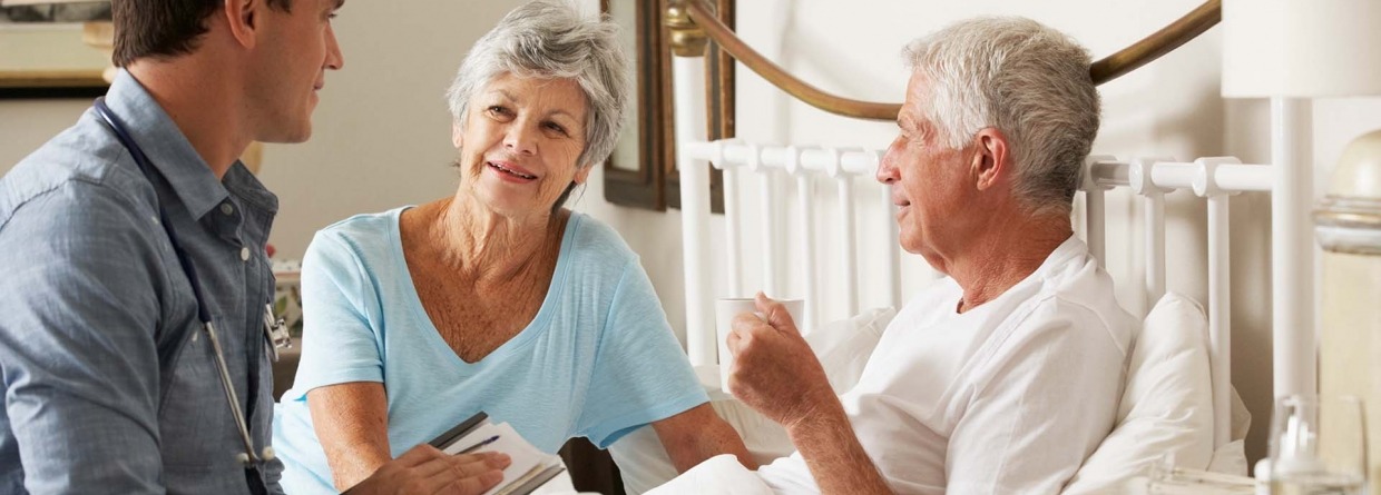 Dokter op huisbezoek bij senioren waarvan de man in bed ligt en de gezondheid van de man wordt besproken met de vrouw